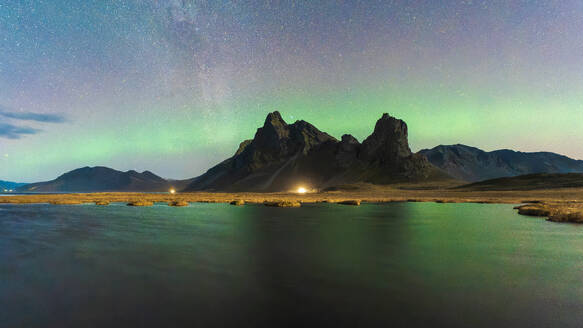 A breathtaking view of the northern lights weaving across a starlit sky above craggy peaks, with a serene lake foreground in Eystrahorn, Iceland - ADSF51571