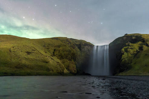 Skogafoss waterfall cascades down a lush cliffside under a captivating night sky tinged with aurora-like lights in Iceland - ADSF51573