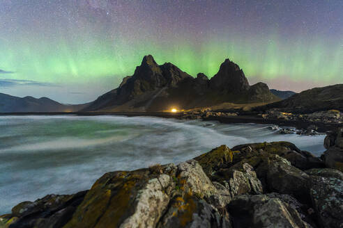 The mesmerizing Northern Lights cascade over a rugged coastline with sharp mountain peaks and a smooth, flowing ocean in Eystrahorn, Iceland - ADSF51576