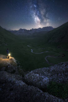 A lone hiker stands atop a mountain, gazing at the Milky Way above a winding river in a valley in Pyrenees, Spain - ADSF51586