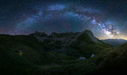 A breathtaking panorama featuring the Milky Way arcing over a tranquil mountain range with a lone hiker with flashlight in Pyrenees, Spain - ADSF51587