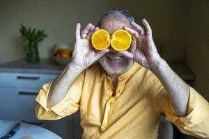 Cheerful senior man in casual clothes smiling and covering eyes with halved orange slices while sitting n kitchen - ADSF51597