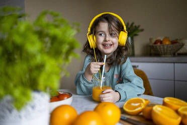 Smiling adorable girl listening to music through headphones and looking away while drinking orange juice on table at home - ADSF51598