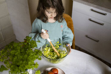 From above elementary cute girl wearing casuals mixing vegetables with spatula in glass bowl while preparing salad at home - ADSF51608