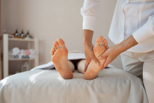 Crop hands of anonymous female therapist massaging foot of customer lying on bed to relieve pain by gently pressing pressure points at spa salon - ADSF51613