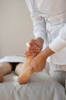 Crop hands of anonymous female therapist massaging foot of customer lying on bed to relieve pain by gently pressing pressure points at spa salon - ADSF51615