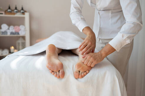 Crop hands of anonymous female therapist massaging foot of customer lying on bed to relieve pain by gently pressing pressure points at spa salon - ADSF51616