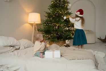 Two children experience the joy of the holiday season next to a beautifully decorated Christmas tree, with one toddler playing with a toy and the other admiring the tree lights. - ADSF51619