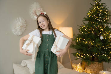 A smiling young girl with reindeer antlers holds Christmas presents, standing in front of a decorated tree with twinkling lights. - ADSF51620