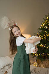 A cheerful young girl wearing reindeer antlers smiles as she holds a wrapped Christmas gift, with a festive tree in the background. - ADSF51621