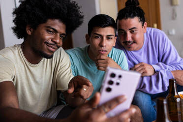 Smiling young man with afro hair taking selfie over smartphone with male friends while spending leisure time together in living room - ADSF51632