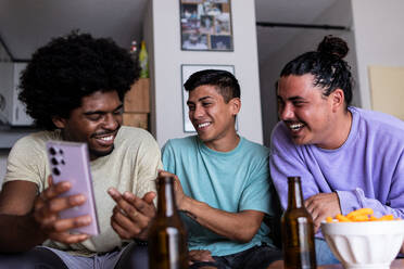 Smiling young man with afro hair showing a photo over smartphone to male friends while spending leisure time together in living room - ADSF51633