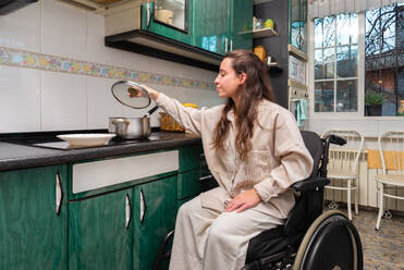 A woman in a wheelchair comfortably reaches for a pot lid while cooking in an adapted kitchen space - ADSF51685