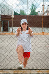 A woman looking at camera in sporty wear stretches her leg on a chain-link fence at an outdoor court - ADSF51692