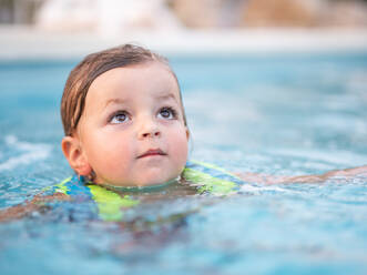 Closeup of little boy wearing inflatable jacket looking up while swimming in pool and enjoying vacation during summer - ADSF51714