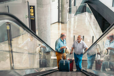 High angle view of senior male travelers in casuals talking with each other while standing with luggage on moving stairs at airport terminal - ADSF51756