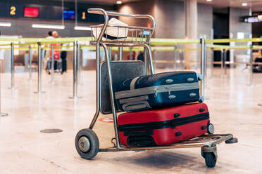 Colorful suitcases with hat placed on steel trolley in modern airport terminal before flight during trip against blurred background - ADSF51757