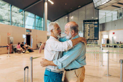 Side view of happy senior male friends dressed in casual clothes embracing while standing with luggage at modern airport terminal during farewell - ADSF51763