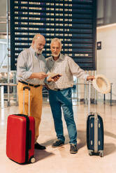 Serious senior male passenger friends in casual clothes checking boarding pass while standing against timetable and waiting for departure at modern airport - ADSF51764