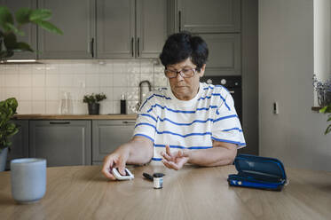 Ältere Frau misst den Blutzucker mit einem Blutzuckermessgerät in der Küche - ALKF00924