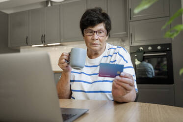 Ältere Frau beim Bezahlen mit der Kreditkarte über einen Laptop in der Küche - ALKF00931