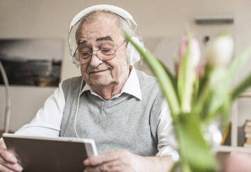 Lächelnder älterer Mann mit Kopfhörern und einem Tablet-PC zu Hause - UUF30857