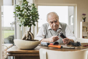 Senior man sitting near window and examining miniature train through magnifying glass at home - UUF30863