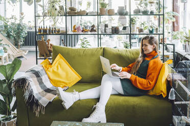 Smiling botanist using laptop on sofa in plant store - OLRF00062