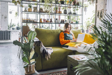 Botanist sitting on sofa and using laptop in plant store - OLRF00066