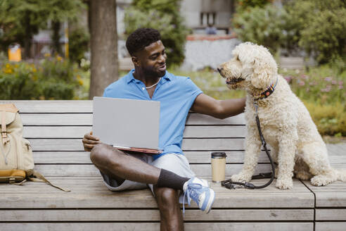 Smiling young man with laptop stroking dog while sitting on bench in park - MASF41422