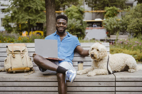 Portrait of smiling man with laptop sitting by labradoodle dog on bench in park - MASF41423