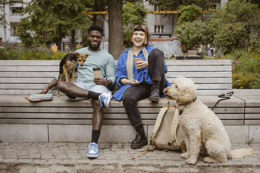 Happy young man and woman holding disposable coffee cups while sitting with dogs in park - MASF41426