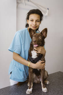 Portrait of female nurse embracing dog on examination table in medical clinic - MASF41493