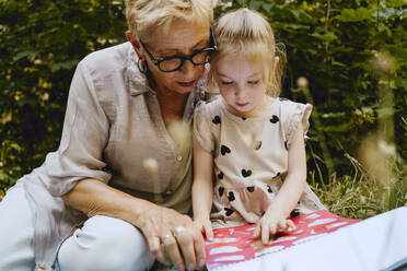 Granddaughter reading book while sitting with grandmother at park - MASF41501