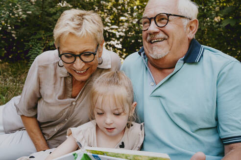 Happy grandparents sitting with granddaughter reading book at park - MASF41502