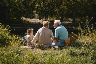 Rear view of grandparents sitting with granddaughter near pond at park - MASF41504