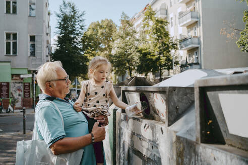 Senior man carrying granddaughter trashing garbage in can at street - MASF41506