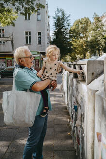 Grandfather carrying daughter trashing garbage while standing at street - MASF41507