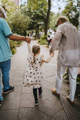 Grandparents holding hands of granddaughter while walking on footpath at street - MASF41524