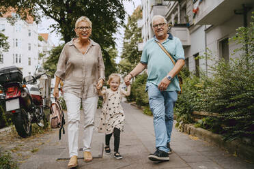 Happy grandparents holding hands of granddaughter while walking at street - MASF41526