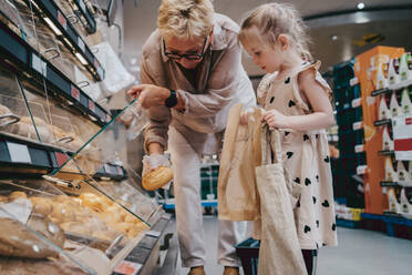 Senior woman buying bread with granddaughter holding bags at grocery store - MASF41529