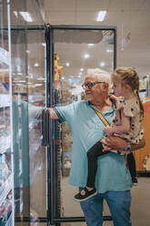 Senior man carrying granddaughter while doing shopping at grocery store - MASF41531