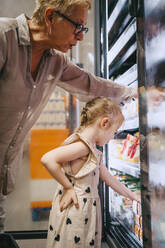 Side view of girl with hand on hip doing shopping from refrigerated section near grandmother at store - MASF41535