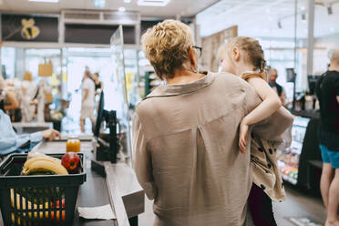 Grandmother carrying granddaughter while standing at checkout counter in supermarket - MASF41539