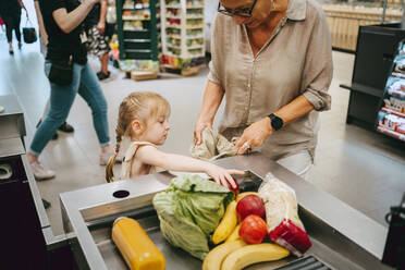 Girl helping grandmother filling groceries in bag at checkout in supermarket - MASF41544