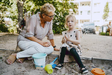 Grandmother talking to granddaughter sitting in sandbox while playing at park - MASF41553