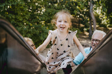 Portrait of smiling girl climbing on slide while playing with grandparents at park - MASF41554