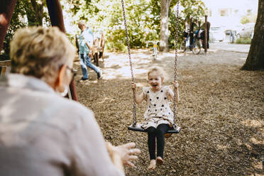 Cheerful girl playing on swing with grandmother at park - MASF41555
