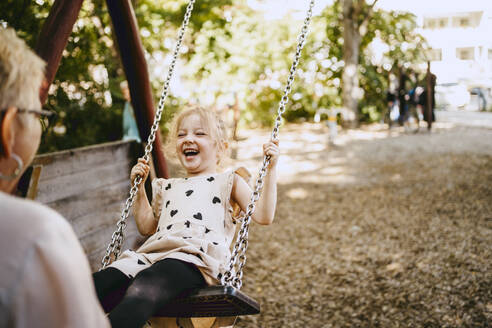 Happy girl laughing while playing on swing with grandmother at park - MASF41556