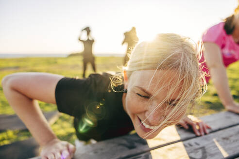 Smiling blond woman doing push-ups on bench during group training at beach - MASF41565